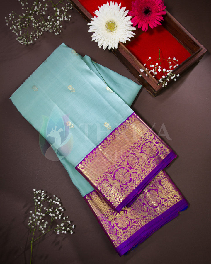 Flat lay of folded Thriya silk saree with pastel blue body and purple zari border, styled with fresh flowers