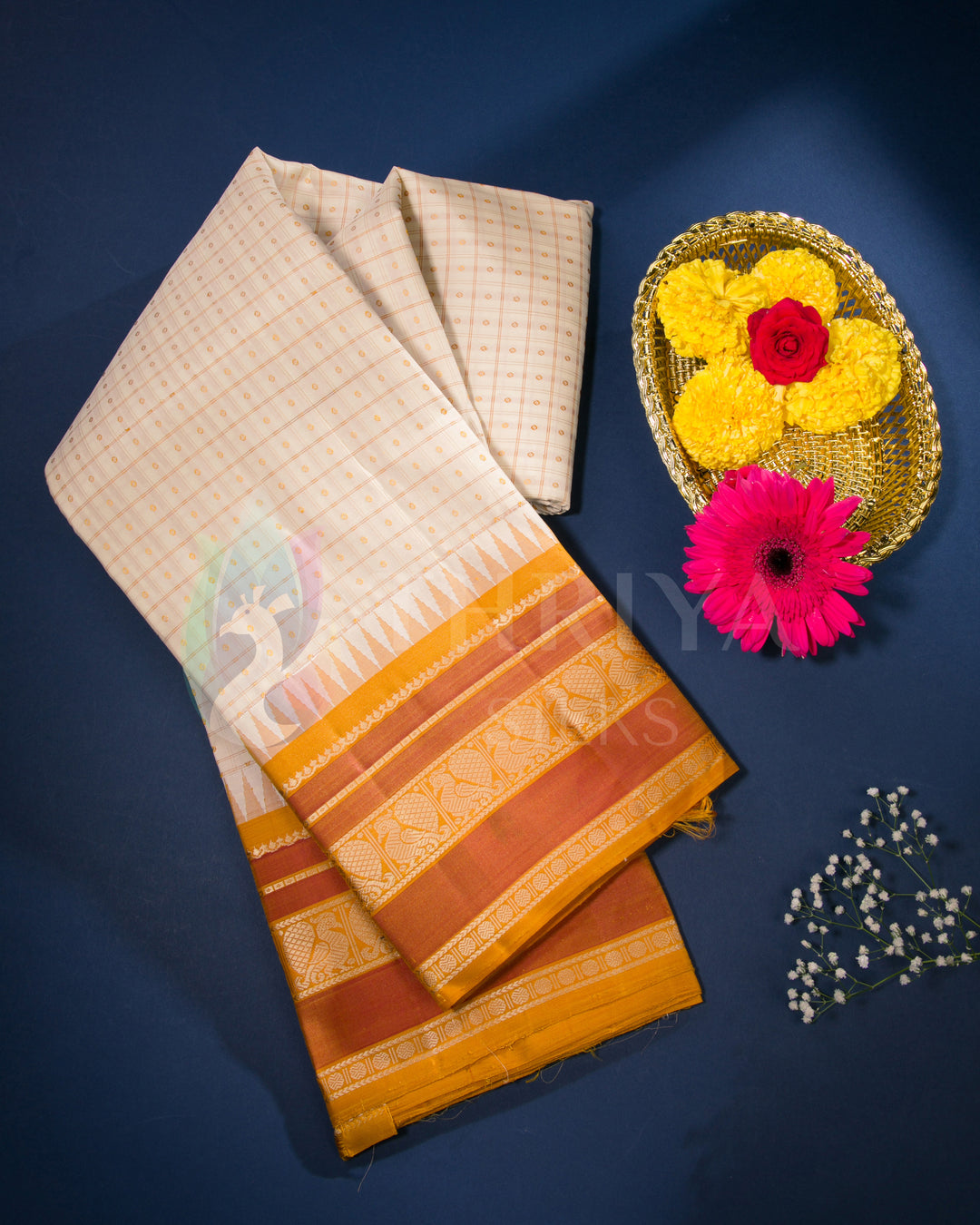 Cream silk saree with orange zari border styled with festive flowers on blue backdrop