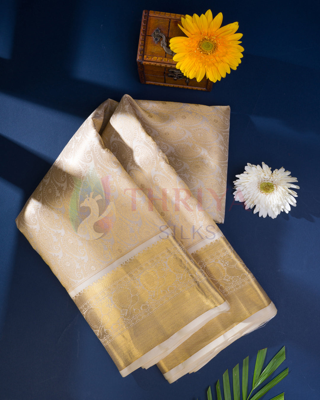 Festive half white Kanchipuram silk saree displayed flat with flowers and a gift box on a blue background, Thriya Silks branding visible
