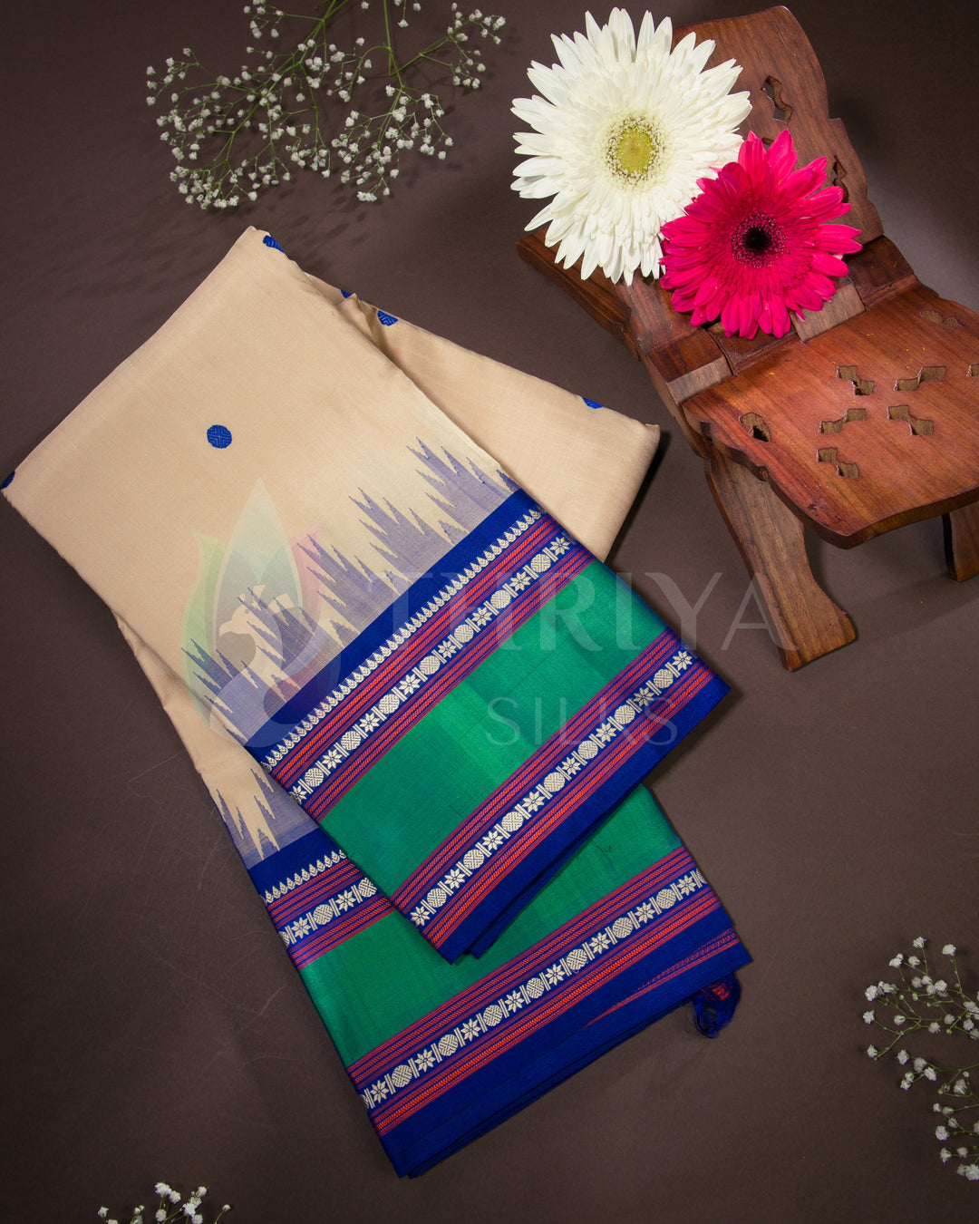 Folded beige silk saree with green and blue zari border, styled with floral decor, from Thriya Silks