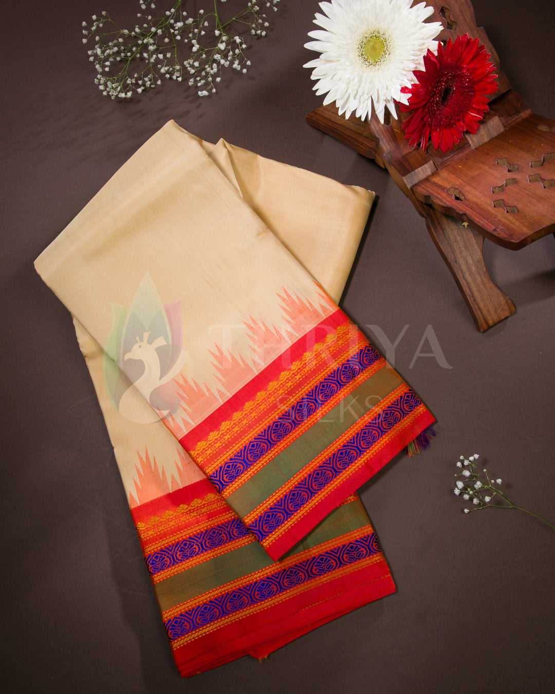 Folded beige silk saree with bright red-orange border and blue zari, styled with floral decor, from Thriya Silks