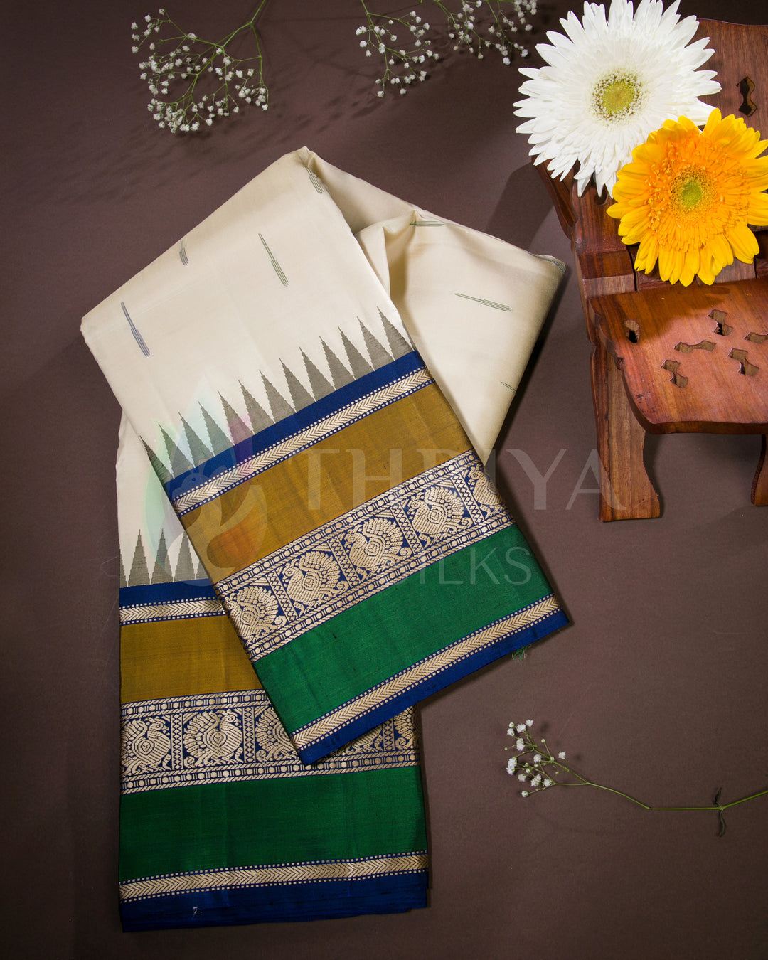 Folded cream silk saree with green, gold, and blue borders in a lifestyle setting with floral décor, from Thriya Silks