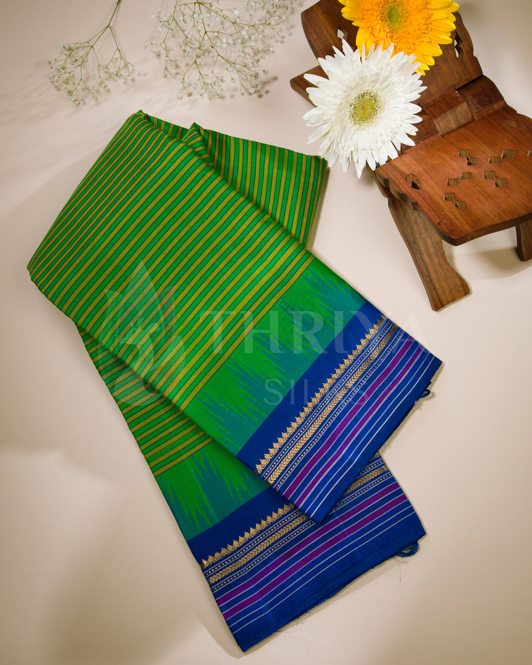 Folded green and blue silk saree displayed with white and yellow flowers, from Thriya Silks
