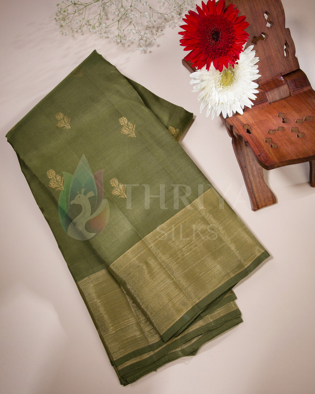 Folded olive green Kanjivaram silk saree with golden zari, styled with flowers on beige background