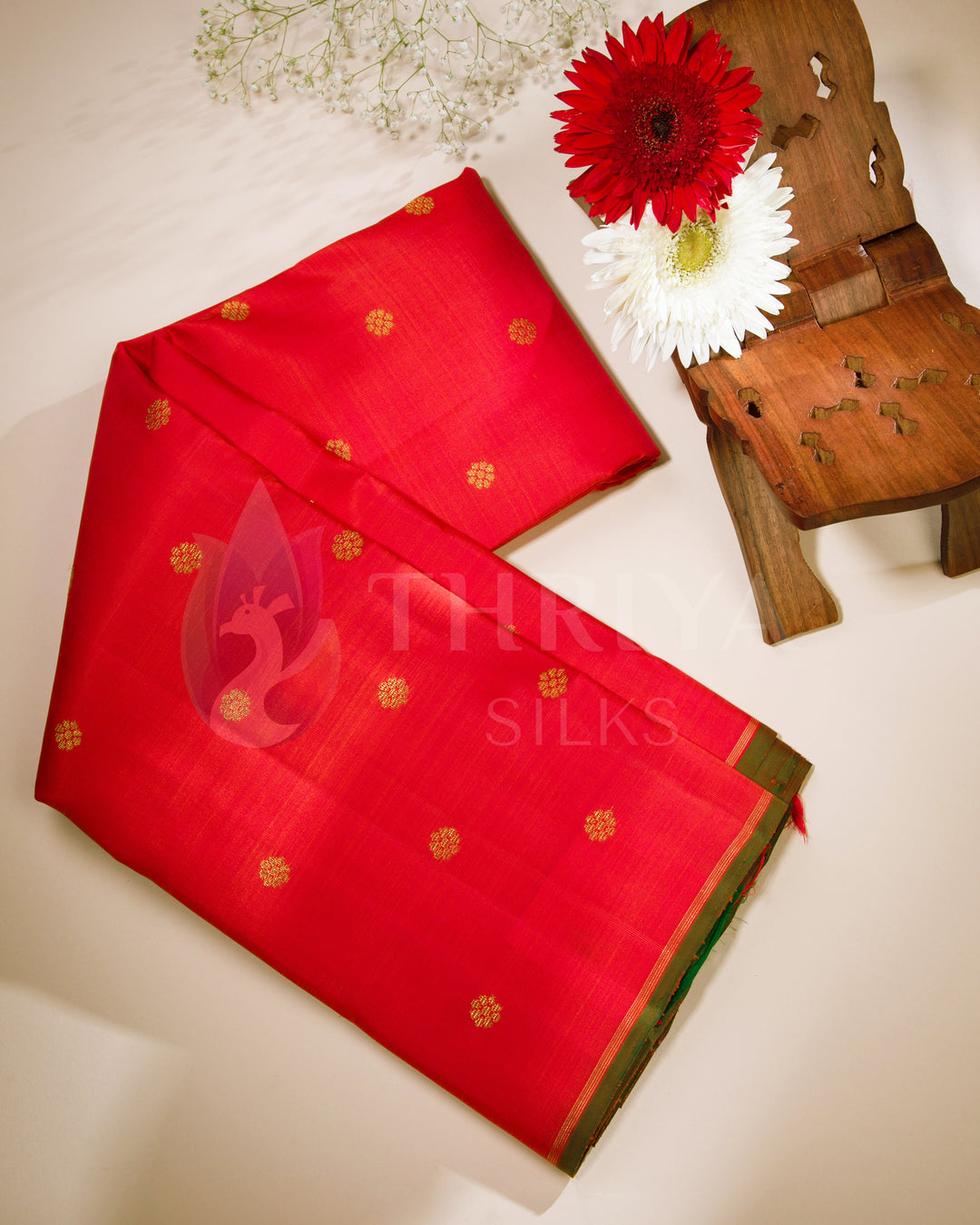 Folded red Kanjivaram silk saree with gold motifs and green edge – lifestyle photo with floral decor from Thriya Silks