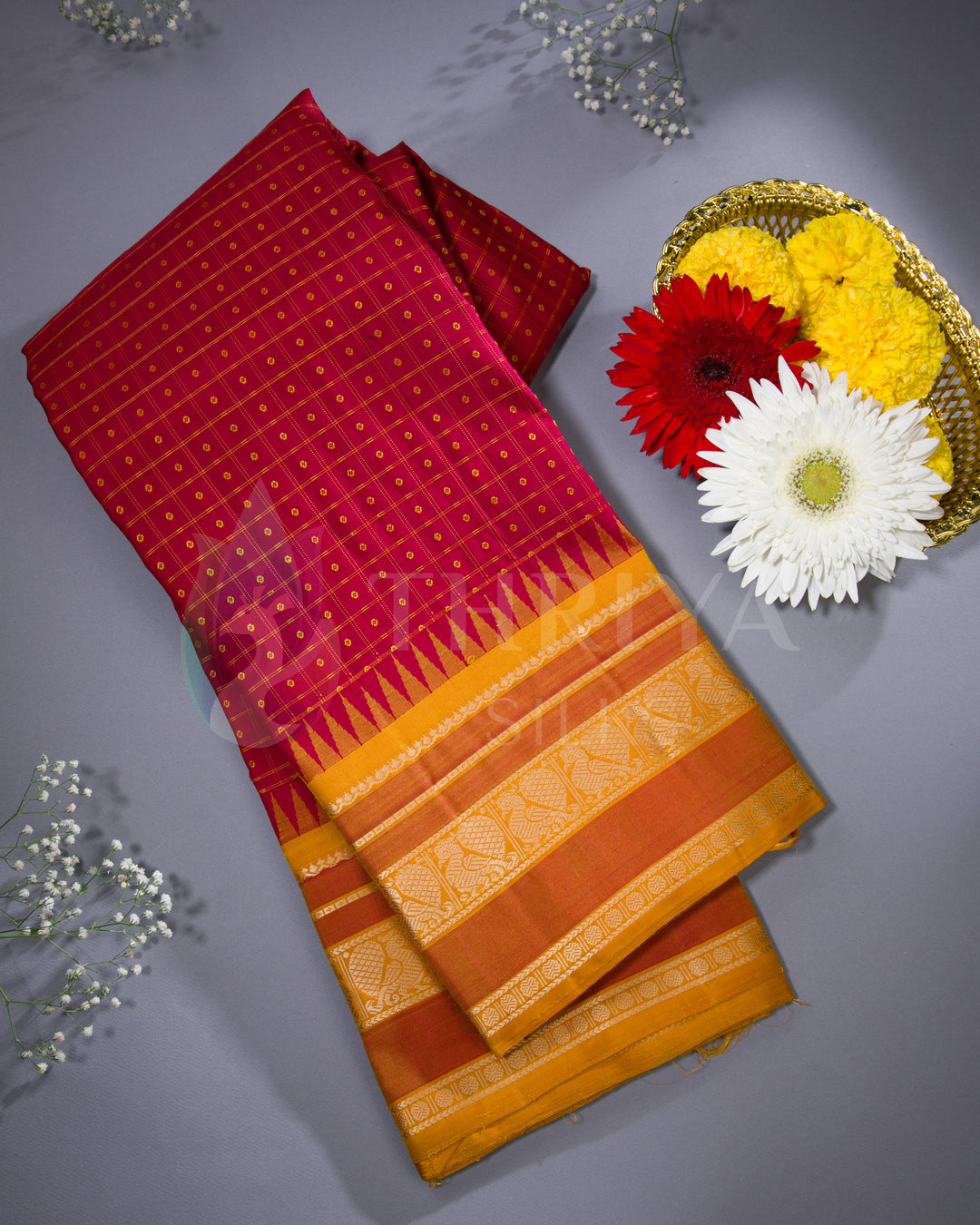 Flatlay of folded red silk saree with mustard yellow zari border, styled with floral decor from Thriya Silks