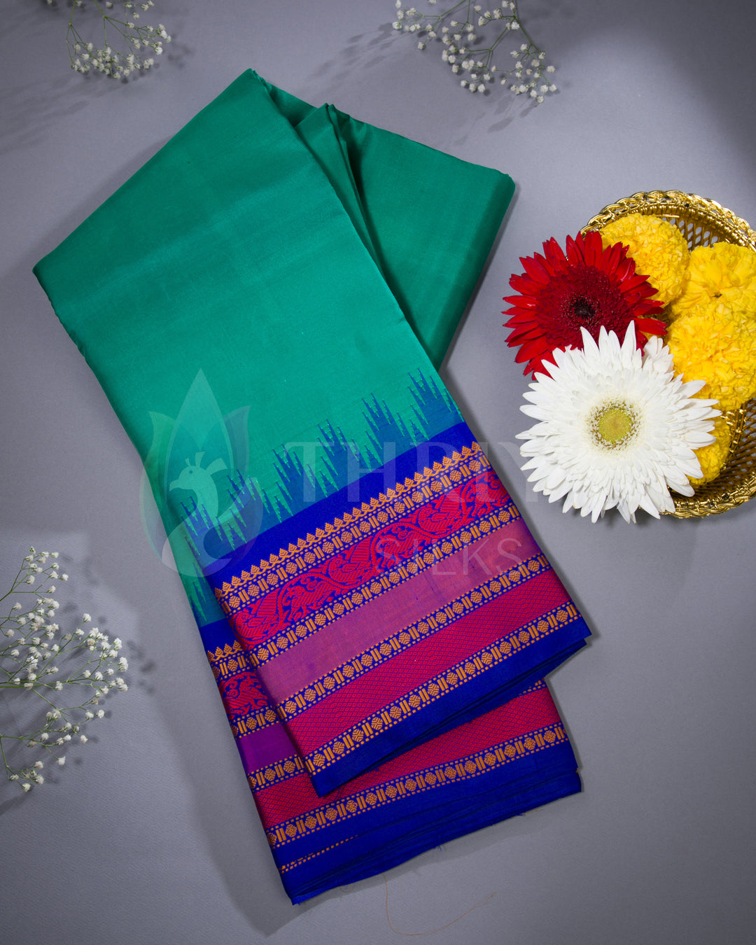 Folded teal green silk saree with royal blue and magenta border – styled flatlay with flowers by Thriya Silks
