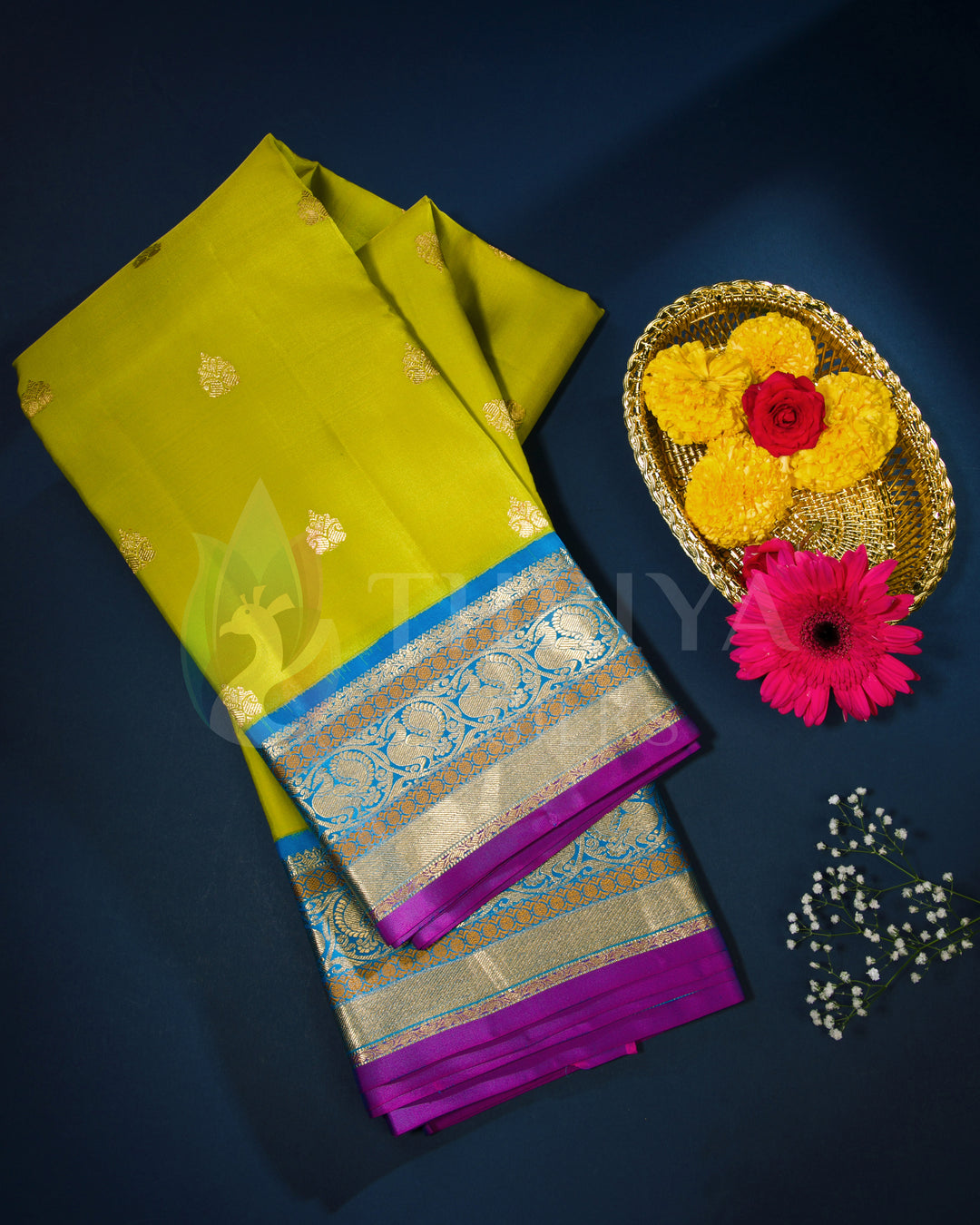 Folded lime yellow silk saree with royal blue border, paired with festive flowers in flat lay style