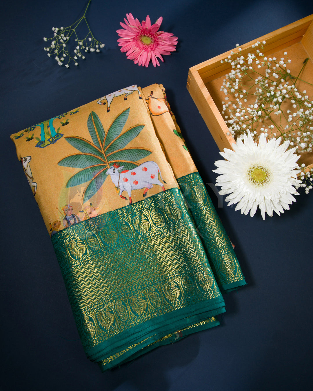 Folded view of mustard yellow silk saree featuring cow and palm tree motifs with a shimmering green zari border