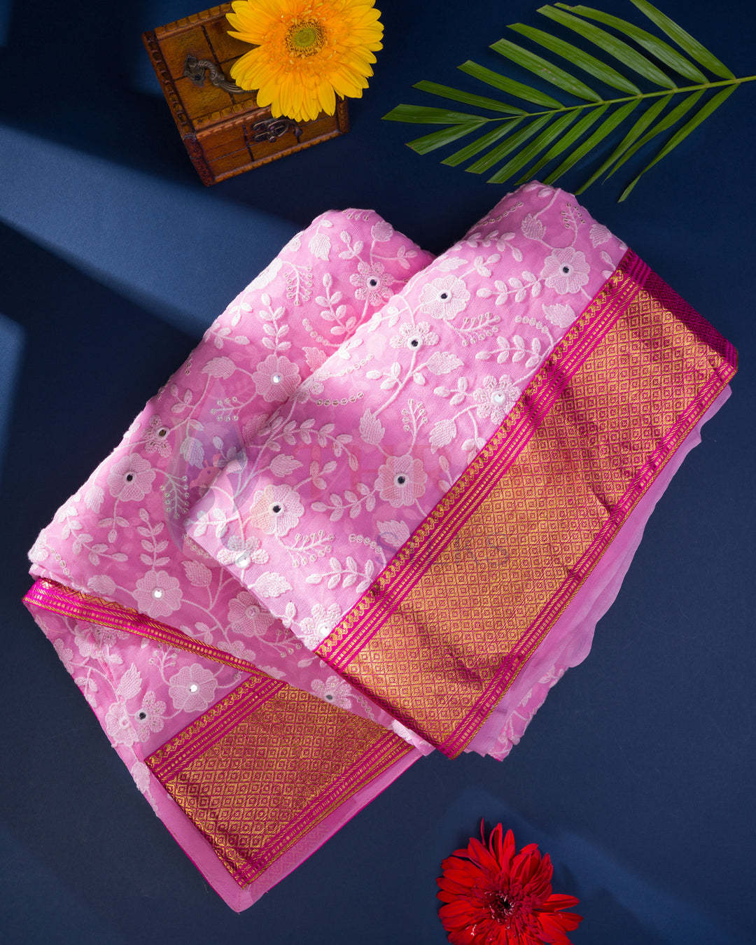 Pink Chikankari silk saree with floral border, styled on a blue surface with flowers
