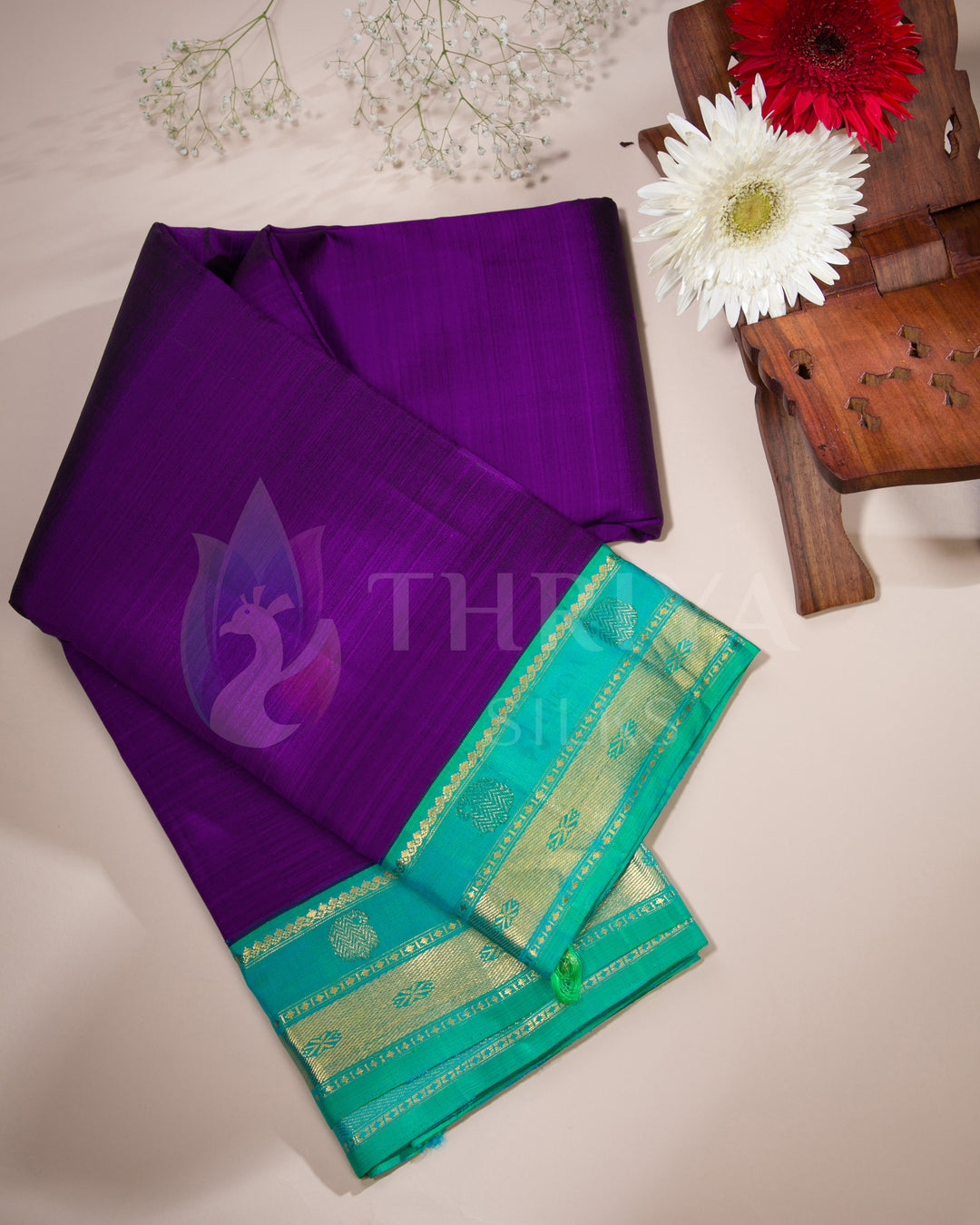 Styled flatlay of purple silk saree with aqua blue zari border placed beside flowers
