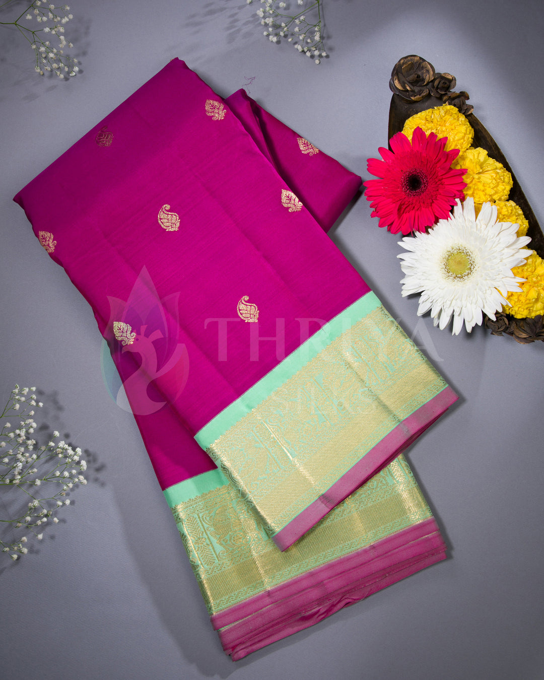 Styled presentation of rani pink silk saree with gold motifs, light green border and pallu, paired with flowers