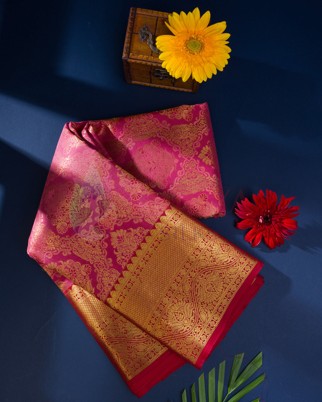 Rust Kanjivaram silk saree folded with gold zari work, styled with flowers and gift box on blue backdrop